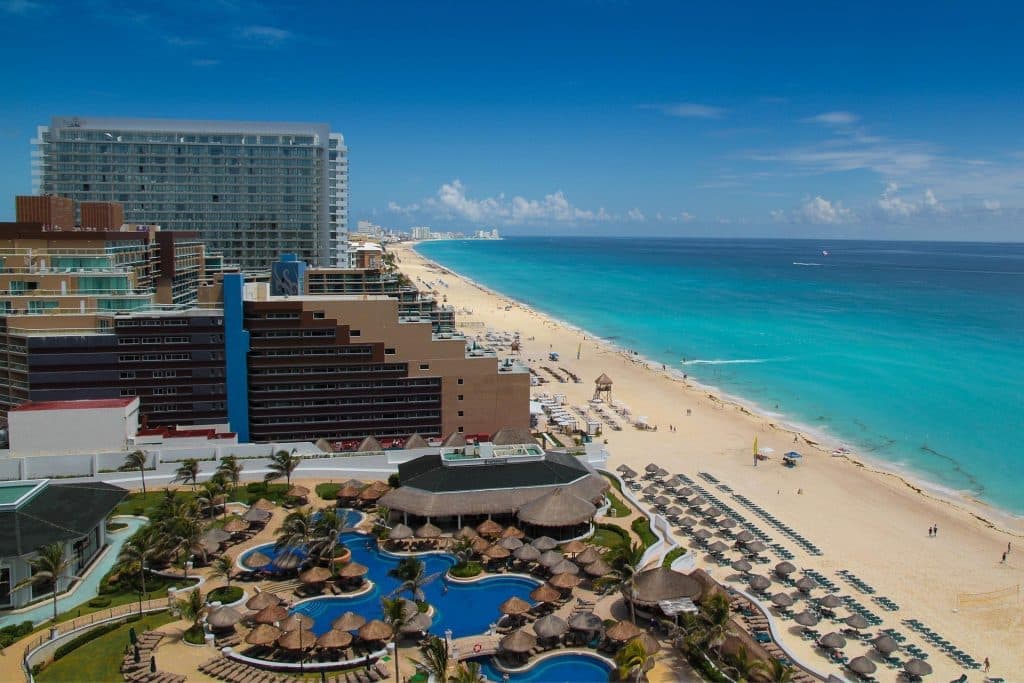 Aerial view of a beachfront resort with pools, palm trees, and lounge chairs, next to a long stretch of white sandy beach and clear turquoise ocean under a bright blue sky in Cancun, Mexico.
