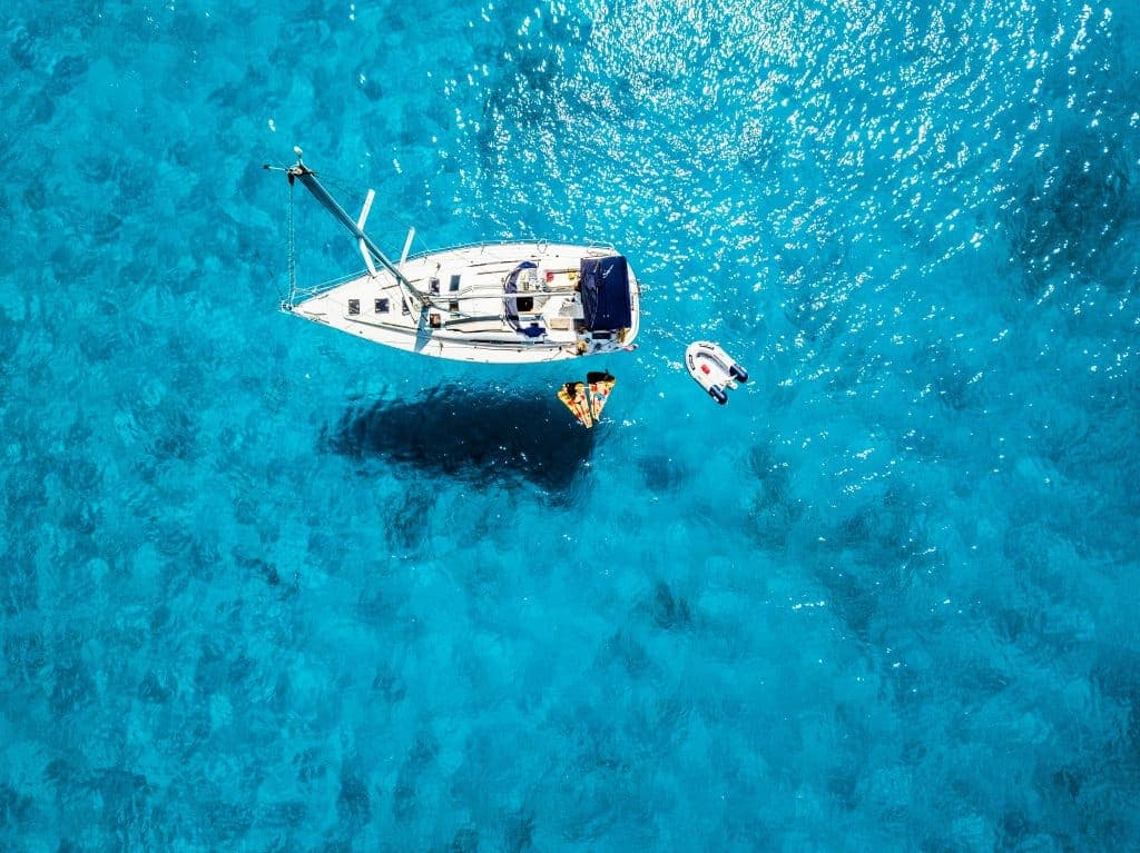 Aerial view of a white sailboat anchored in clear blue water with two people relaxing on orange floats beside it and a small inflatable boat nearby. Sunlight sparkles on the surface of the sea.