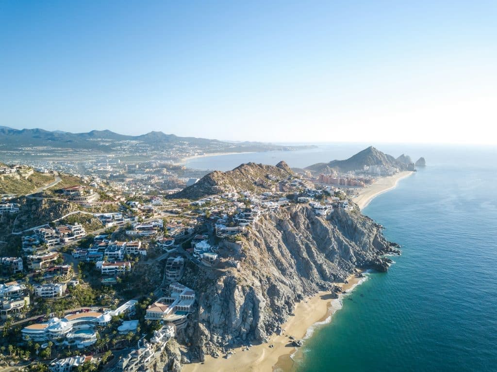Aerial view of a coastal landscape featuring rugged cliffs and sandy beaches. Buildings dot the cliffside, leading to a serene ocean. Mountains are visible in the background under a clear blue sky.