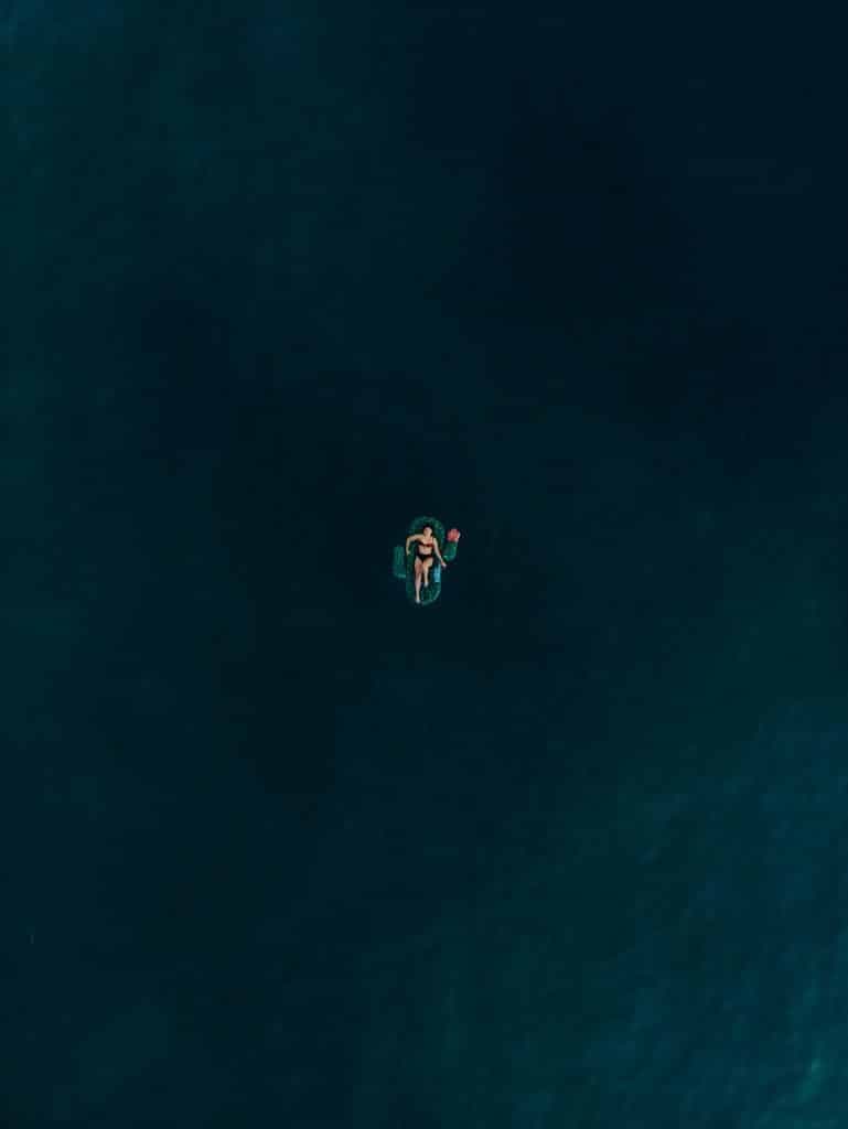 A person floats on their back in deep, dark water. They are centered in the image, surrounded by a vast expanse of water, creating a sense of isolation and tranquility.