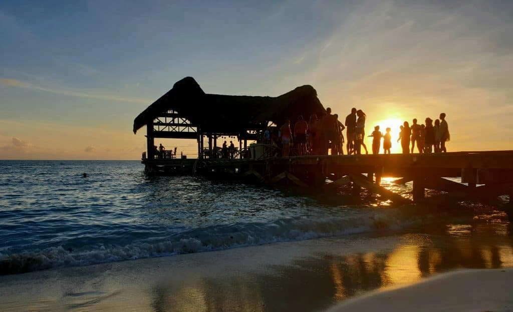 Silhouetted people gather on a pier with a thatched-roof hut, watching the sunset over calm ocean waves; golden sunlight reflects on wet sand and water.