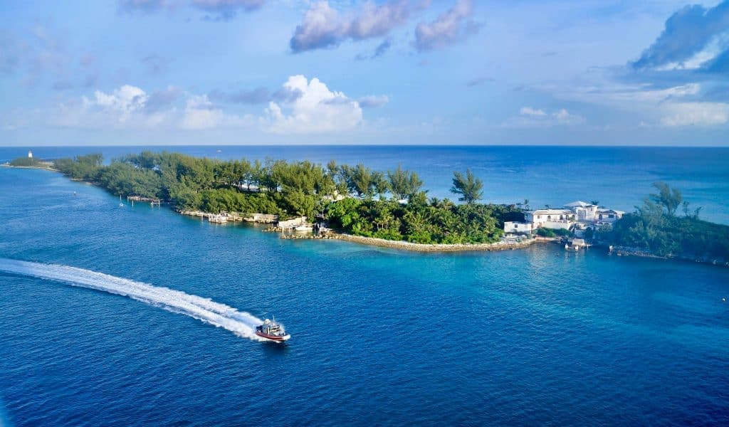A small boat speeds across vibrant blue water near a lush, green island with white buildings and dense trees, under a partly cloudy sky.