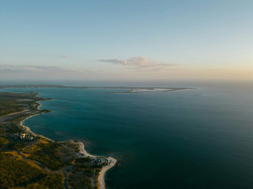 Aerial view of a coastline with clear blue-green water, sandy beaches, and patches of green vegetation, taken at sunset with soft, golden light and a partly cloudy sky.